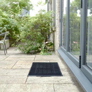 May include: A black doormat with a textured surface sits in front of a glass door leading to a patio. The doormat is made of rubber and has a honeycomb pattern.
