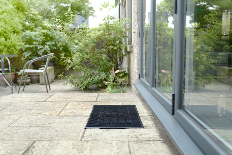 May include: A black doormat with a textured surface sits in front of a glass door leading to a patio. The doormat is made of rubber and has a honeycomb pattern.