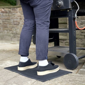 May include: A black grill mat with a person standing on it, wearing black pants and black shoes. The grill mat is in front of a black grill with a green propane tank.