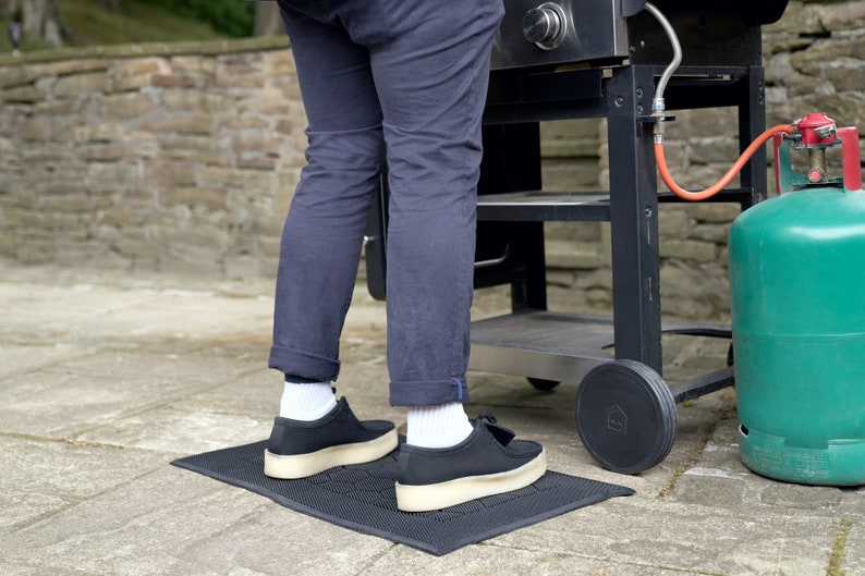 May include: A black grill mat with a person standing on it, wearing black pants and black shoes. The grill mat is in front of a black grill with a green propane tank.