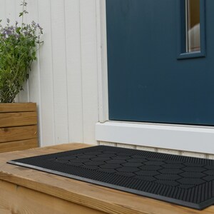 May include: Black doormat with a hexagonal pattern, placed on a wooden porch in front of a blue door.