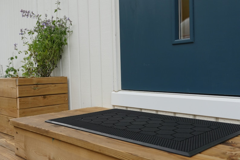 May include: Black doormat with a hexagonal pattern, placed on a wooden porch in front of a blue door.