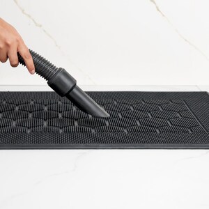 May include: A person uses a vacuum cleaner to clean a black rubber doormat with a honeycomb pattern.