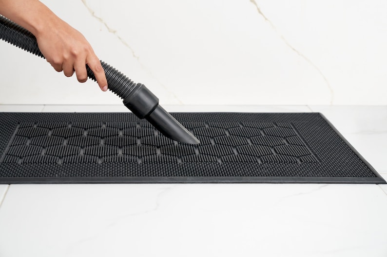 May include: A person uses a vacuum cleaner to clean a black rubber doormat with a honeycomb pattern.