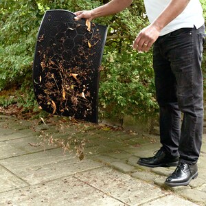 May include: A person is holding a black doormat with brown leaves and debris on it. The doormat is being shaken out over a concrete patio.