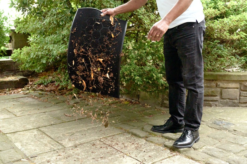 May include: A person is holding a black doormat with brown leaves and debris on it. The doormat is being shaken out over a concrete patio.