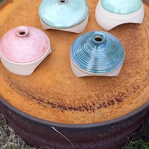 May include: Four ceramic vessels with lids in shades of pink, blue, and green sit on a rusty metal surface. The vessels have a rounded shape with a small opening at the top. The background includes grass and a wooden structure.