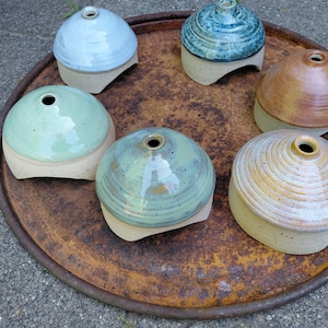 May include: Six dome-shaped ceramic vessels with small circular openings at the top. Each piece features a smooth, glazed upper section in various colours including light blue, teal, green, and tan, contrasted with a speckled, unglazed base.