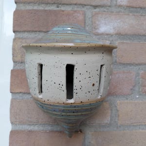 Könnte beinhalten: Ein wandmontiertes Vogelhaus aus Keramik mit einem gesprenkelten, cremefarbenen Körper und vertikalen, rechteckigen Öffnungen. Die obere Kuppel und die spitz zulaufende Basis sind mit einer glatten, glänzenden blaugrauen Glasur versehen.