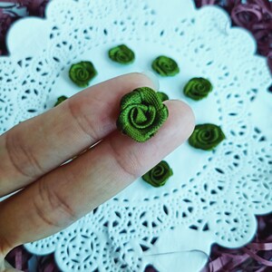 May include: A collection of small, green fabric rose embellishments. The roses are arranged on a white doily with a lace pattern.