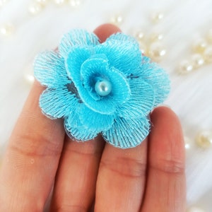以下が含まれることがあります： 真珠の中心を持つターコイズブルーの花のクローズアップ。花は繊細で重ねられた布の花びらでできています。花は手で持たれており、白い布と散らばった真珠のぼやけた背景があります。