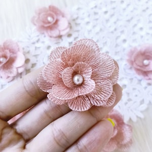 Könnte beinhalten: Nahaufnahme einer rosa Stoffblume mit Perlenmitte. Die Blume ist mehrlagig und hat eine zarte, gekräuselte Textur.