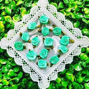 May include: A collection of 15 teal blue satin ribbon rosettes with green stems, arranged on a white lace doily.
