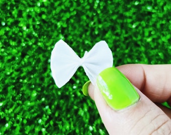 Puede incluir: Un lazo de tela blanco con un nudo central, sostenido por un dedo con esmalte de uñas verde brillante sobre un fondo verde.