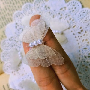 Puede incluir: Un delicado adorno blanco en forma de mariposa con detalles de perlas. El adorno está hecho de tela transparente y es perfecto para agregar un toque de elegancia a las manualidades y proyectos.