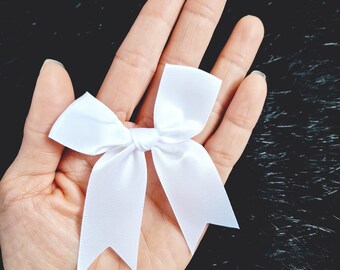 Puede incluir: Un lazo de satén blanco con un nudo central, sostenido en una mano sobre un fondo negro.