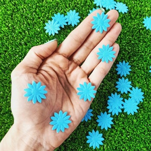May include: A hand holding a variety of light blue felt flower embellishments. The flowers are scattered on a green surface.