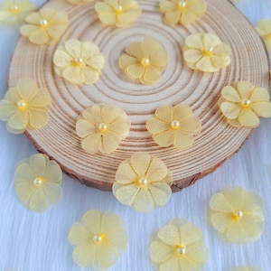 Può includere: Una collezione di fiori di tessuto giallo con centri di perle, disposti su una superficie di legno. I fiori sono sparsi intorno alla superficie, alcuni si sovrappongono.