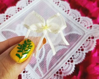 May include: A white sheer bow with a delicate texture, held by a finger with yellow nail polish and green leaf nail art.
