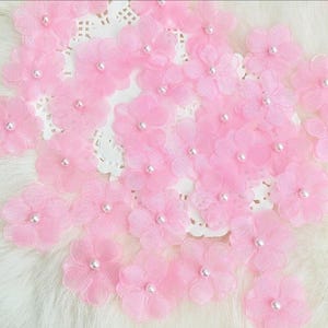 Könnte beinhalten: Eine Sammlung zarter, hellrosa Stoffblumen mit einem Perlenmittelpunkt. Die Blumen sind auf einer weißen, spitzenartigen Oberfläche angeordnet. Diese floralen Verzierungen sind ideal für Bastelprojekte.