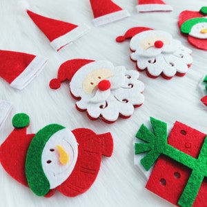 May include: A selection of Christmas felt decorations, featuring Santa Claus faces, snowman shapes, and red Santa hats with white edging. The ornaments are red, white, green, and yellow, displayed on a white, fluffy background.