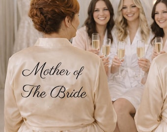 May include: Two women wearing champagne-colored satin robes with the text "Mother of The Bride" and "Mother of The Groom" on the back. The women are surrounded by other women, some holding champagne flutes. The scene suggests a wedding celebration.