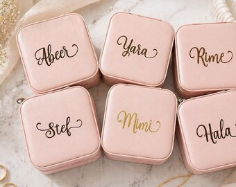 May include: Six square, light pink jewelry cases with personalized names in elegant script: Abeer, Yara, Rime, Stef, Mimi, and Hala. The cases are arranged on a soft, light-colored surface, with gold jewelry and floral accents.