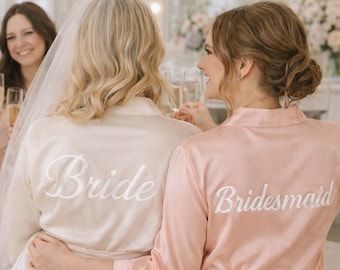 May include: A group of women in a room, celebrating. The bride wears a cream-colored robe with "Bride" embroidered on the back. A bridesmaid wears a pink robe with "Bridesmaid" on the back. They are toasting with champagne flutes.