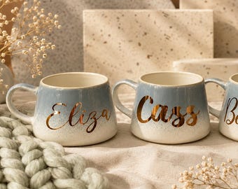 May include: Three ceramic mugs with a blue and white ombre design. Each mug has a gold script name: Eliza, Cass, and Belinda. The mugs are on a neutral-toned surface with decorative elements in the background.