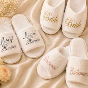 May include: White plush slippers with open toes, embroidered with "Bride", "Maid of Honour", and "Bridesmaid" in gold, black, and pink lettering. The slippers are arranged on a cream-coloured fabric, with floral accents.