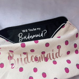 May include: A white canvas zippered pouch with pink polka dots. The pouch is open and the inside lining is black. The text "Will You be my Bridesmaid?" is printed in white on the black lining. The text "Bridesmaid" is printed in pink on the front of the pouch.