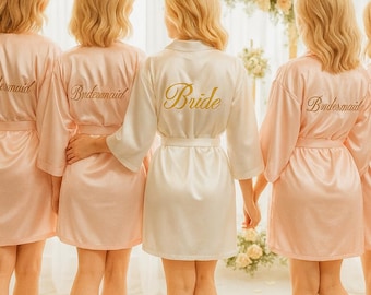 May include: Five women wearing satin robes, four in blush pink with "Bridesmaid" embroidered on the back, and one in white with "Bride" on the back. The group stands in front of a floral arch.