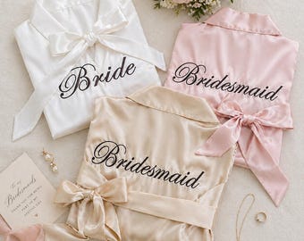May include: Three satin robes in white, pink, and champagne, each with a matching bow and the word "Bride" or "Bridesmaid" embroidered in black script. A bouquet of flowers and jewelry are also visible.