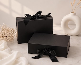 May include: Two black gift boxes with satin ribbon bows. One box is taller than the other. A white vase with a circular opening and dried stems is in the background. The boxes are on a marble surface.