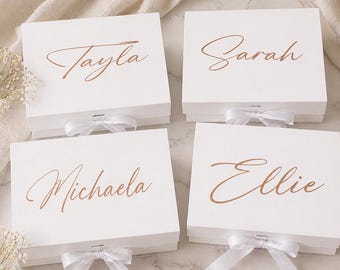 May include: Four white gift boxes with names Tayla, Sarah, Michaela, and Ellie in a copper script. Each box has a white ribbon and bow. The boxes are on a neutral background.