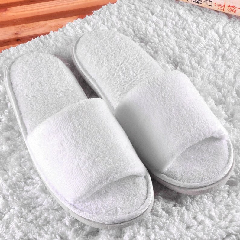 May include: Two white open-toe spa slippers. The slippers are made from a soft, absorbent material and have a simple design. They are positioned on a white, textured surface, suggesting comfort and relaxation. The slippers are ideal for use in a spa or at home.