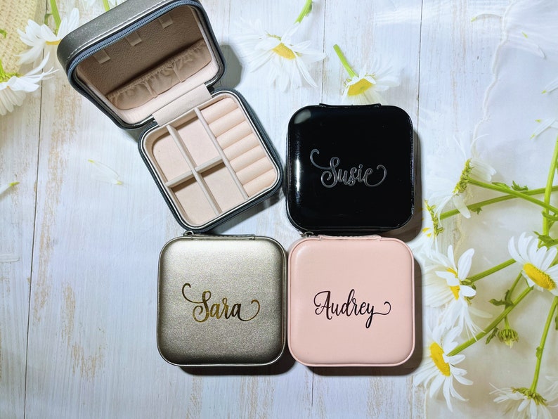 Pu&ograve; includere: Una collezione di portagioie personalizzati in nero, oro e rosa. Ogni custodia quadrata presenta un nome in elegante calligrafia: Susie, Sara e Audrey. Una custodia &egrave; aperta, rivelando scomparti.