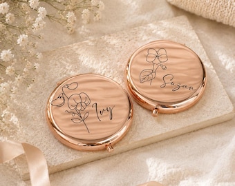 May include: Two rose gold compact mirrors with floral line art and personalized names, "Ivy" and "Suzan". The mirrors are on a light-colored stone surface, with a ribbon and flowers in the background.