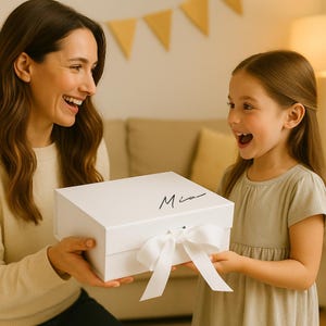 May include: A white gift box with a matching white ribbon and the name "Mia" in black script. The box is being presented to a young girl. The background includes a beige sofa and decorative yellow triangle banners.