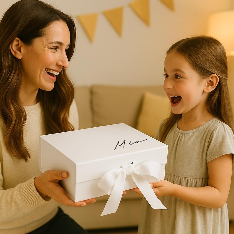 May include: A white gift box with a matching white ribbon and the name "Mia" in black script. The box is being presented to a young girl. The background includes a beige sofa and decorative yellow triangle banners.