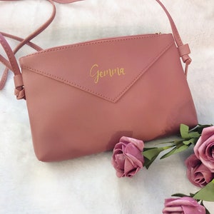 May include: A dusty rose-colored crossbody bag with a gold zipper and the name "Gemma" written in gold script on the front.