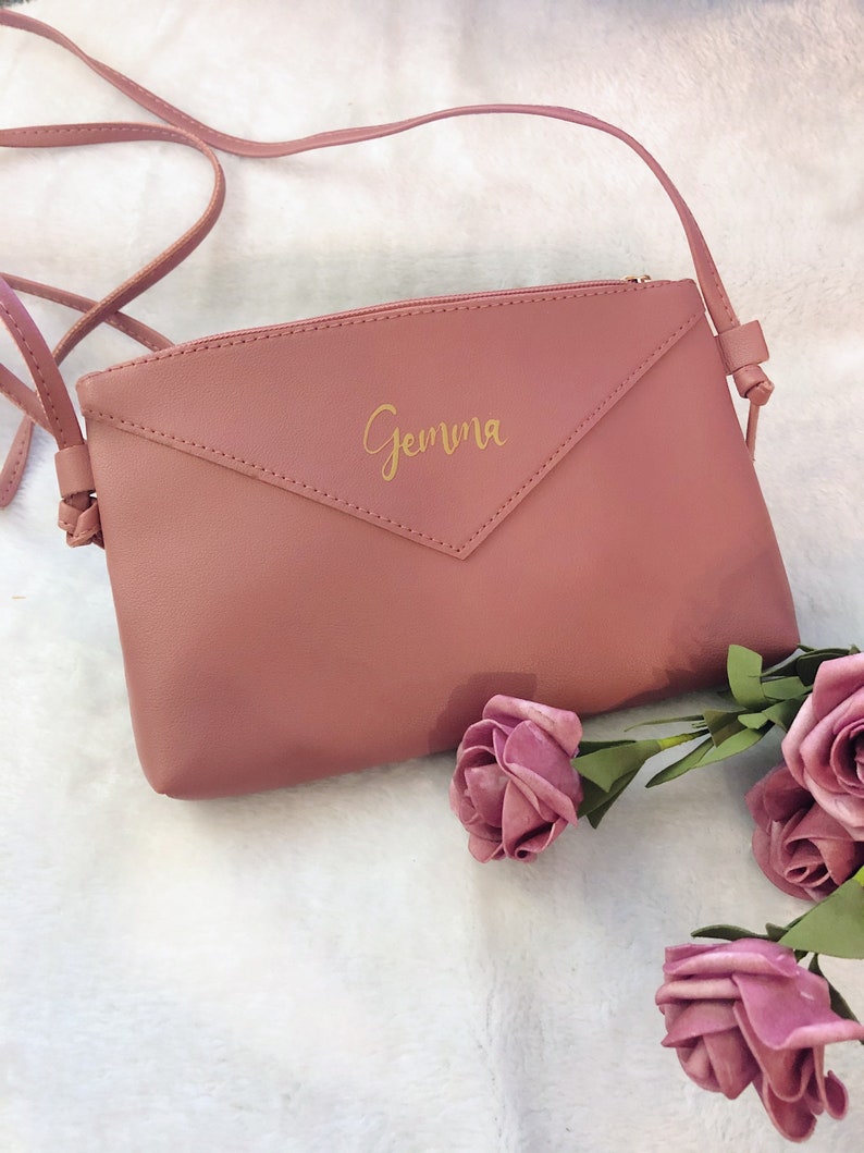 May include: A dusty rose-colored crossbody bag with a gold zipper and the name "Gemma" written in gold script on the front.
