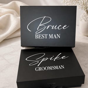 May include: Two black gift boxes with white text. The top box reads "Bruce BEST MAN" in a script font. The bottom box reads "Spike GROOMSMAN" in a script font and has a black ribbon tied around it. The boxes are on a light-colored surface.