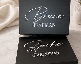 May include: Two black gift boxes with white text. The top box reads "Bruce BEST MAN" in a script font. The bottom box reads "Spike GROOMSMAN" in a script font and has a black ribbon tied around it. The boxes are on a light-colored surface.