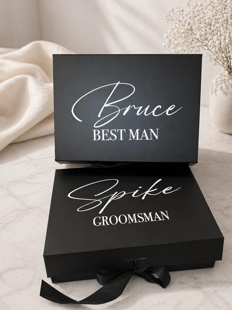 May include: Two black gift boxes with white text. The top box reads "Bruce BEST MAN" in a script font. The bottom box reads "Spike GROOMSMAN" in a script font and has a black ribbon tied around it. The boxes are on a light-colored surface.