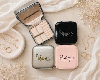 May include: An open, gray jewelry travel case with a beige interior sits above three closed, square jewelry cases. The closed cases are black, silver, and pink, each with a name in cursive. Gold necklaces and rings are also visible.