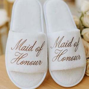 May include: Two white, fluffy slippers embroidered with the words "Maid of Honour" in brown script. The slippers are placed on a light wooden surface, with a blurred background of flowers and soft textures.