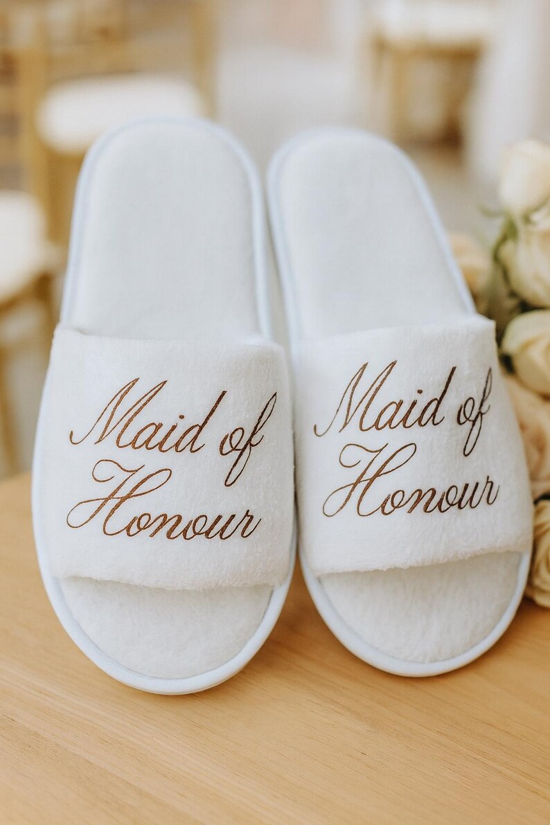 May include: Two white, fluffy slippers embroidered with the words "Maid of Honour" in brown script. The slippers are placed on a light wooden surface, with a blurred background of flowers and soft textures.