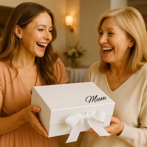 May include: A white gift box with a white bow and the word "Mum" in cursive. Two women are smiling and holding the box. The box is rectangular and has a lid. The bow is tied in the center of the box.