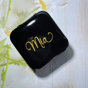 Pu&ograve; includere: Un portagioie nero, quadrato e lucido con il nome "Mia" in oro. La scatola &egrave; chiusa e poggia su uno sfondo floreale bianco e verde chiaro con fiori di margherita.